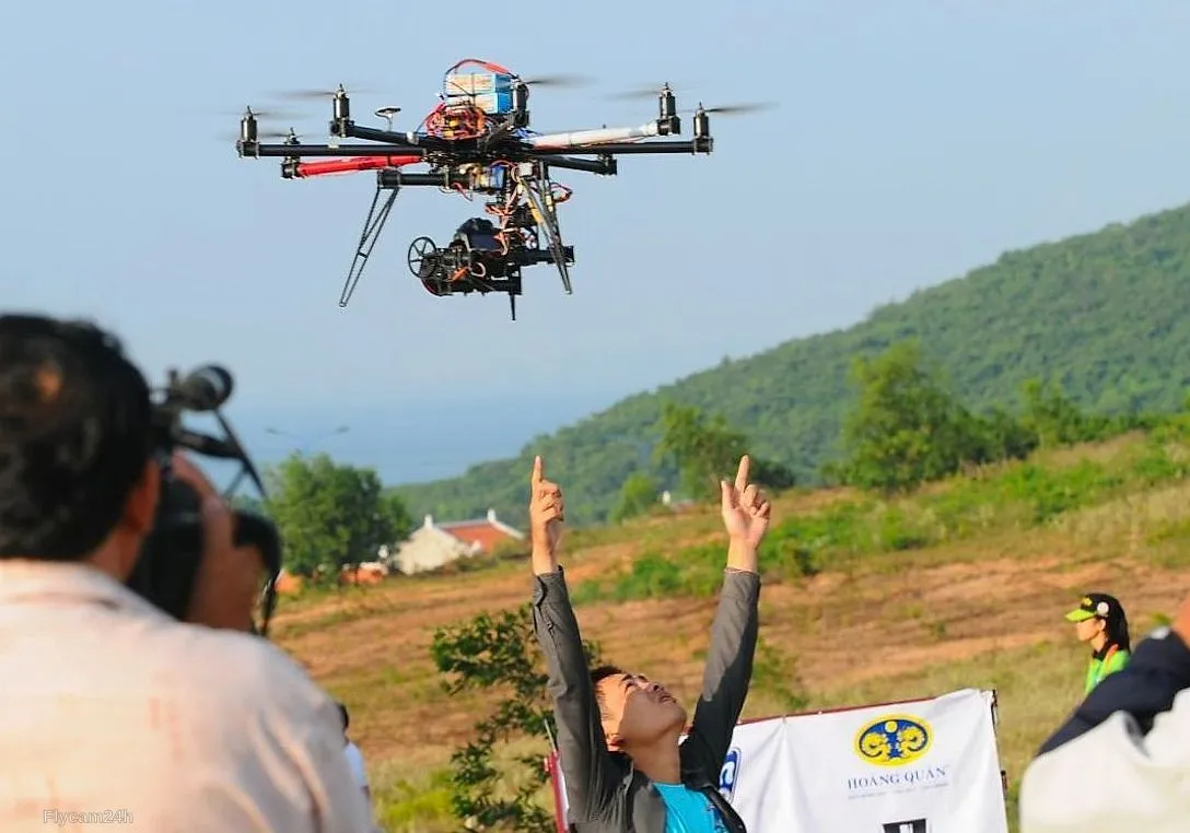 Flycam sáu cánh mang máy quay lớn bay trên sườn đồi tại một sự kiện ngoài trời, người phía dưới giơ tay ra hiệu