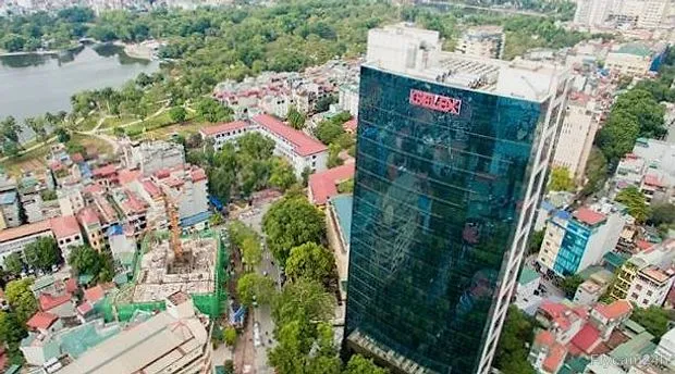Tòa nhà văn phòng mặt kính cao tầng giữa khu phố thấp tầng Hà Nội, phía sau là hồ nước và cây xanh, nhìn chéo từ flycam