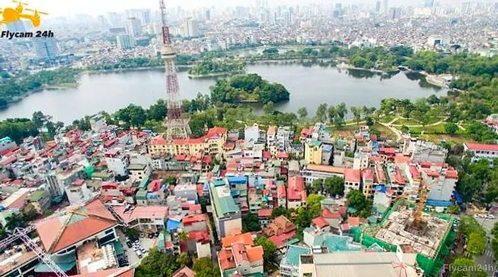 Công trình phần thô có cẩu tháp và lưới che xanh giữa khu dân cư Hà Nội, phía xa là hồ và tháp truyền hình, nhìn chéo từ flycam