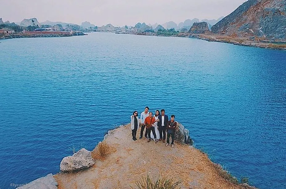 Nhóm bạn đứng trên mỏm đất nhô ra hồ nước xanh, xa xa là núi đá vôi, nhìn từ flycam