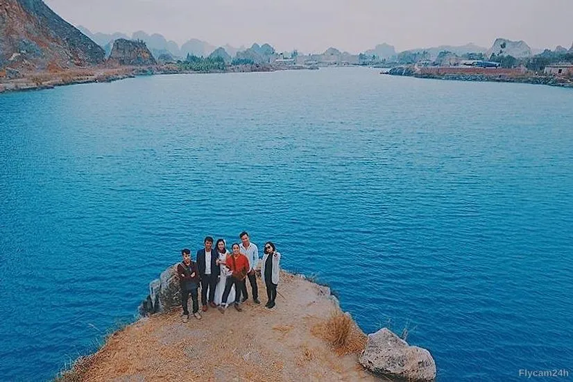Nhóm bạn đứng trên mỏm đất nhô ra hồ nước xanh, xa xa là núi đá vôi, nhìn từ flycam