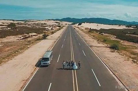 Cô dâu chú rể và nhóm bạn đứng giữa con đường xuyên đồi cát, cạnh chiếc xe khách, nhìn từ flycam