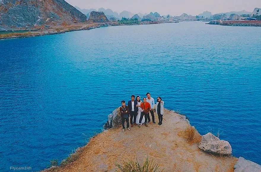 Nhóm bạn đứng trên mỏm đất nhô ra hồ nước xanh, xa xa là núi đá vôi, nhìn từ flycam