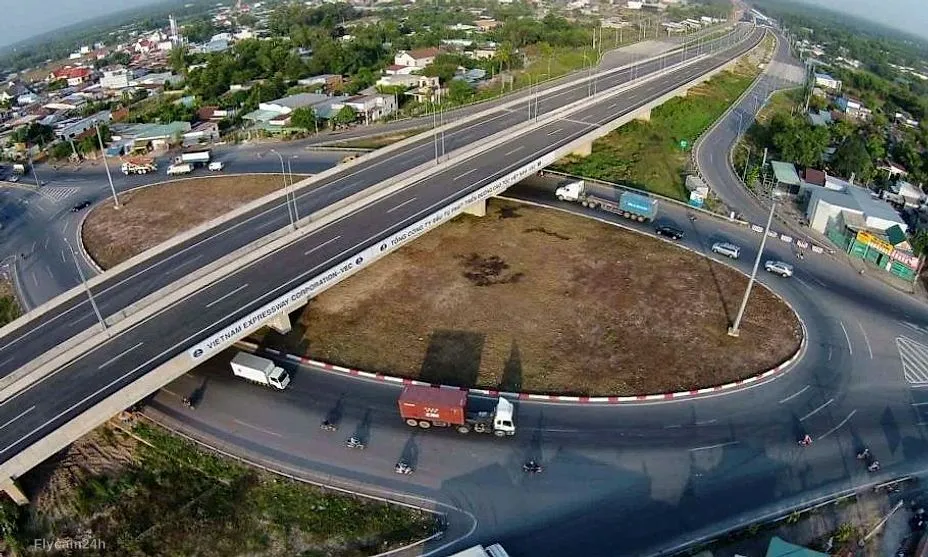 Nút giao cao tốc có cầu vượt và đường vòng, xe tải lưu thông, khu dân cư hai bên, nhìn chéo từ flycam