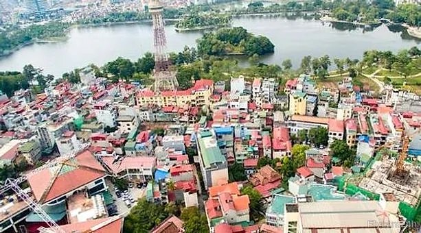 Công trình phần thô có cẩu tháp và lưới che xanh giữa khu dân cư Hà Nội, phía xa là hồ và tháp truyền hình, nhìn chéo từ flycam
