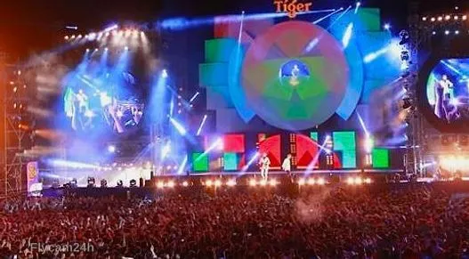 Sân khấu ca nhạc ngoài trời về đêm với màn LED lớn, dàn đèn và đám đông khán giả kín sân, nhìn từ trên cao