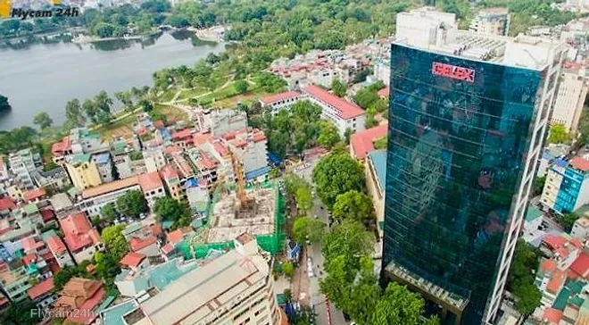 Tòa nhà văn phòng mặt kính cao tầng giữa khu phố thấp tầng Hà Nội, phía sau là hồ nước và cây xanh, nhìn chéo từ flycam