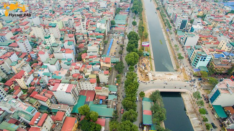 Đoạn kênh trong phố đang thi công cầu và bờ kè, hai bên là đường và nhà dân, chụp chéo từ trên cao